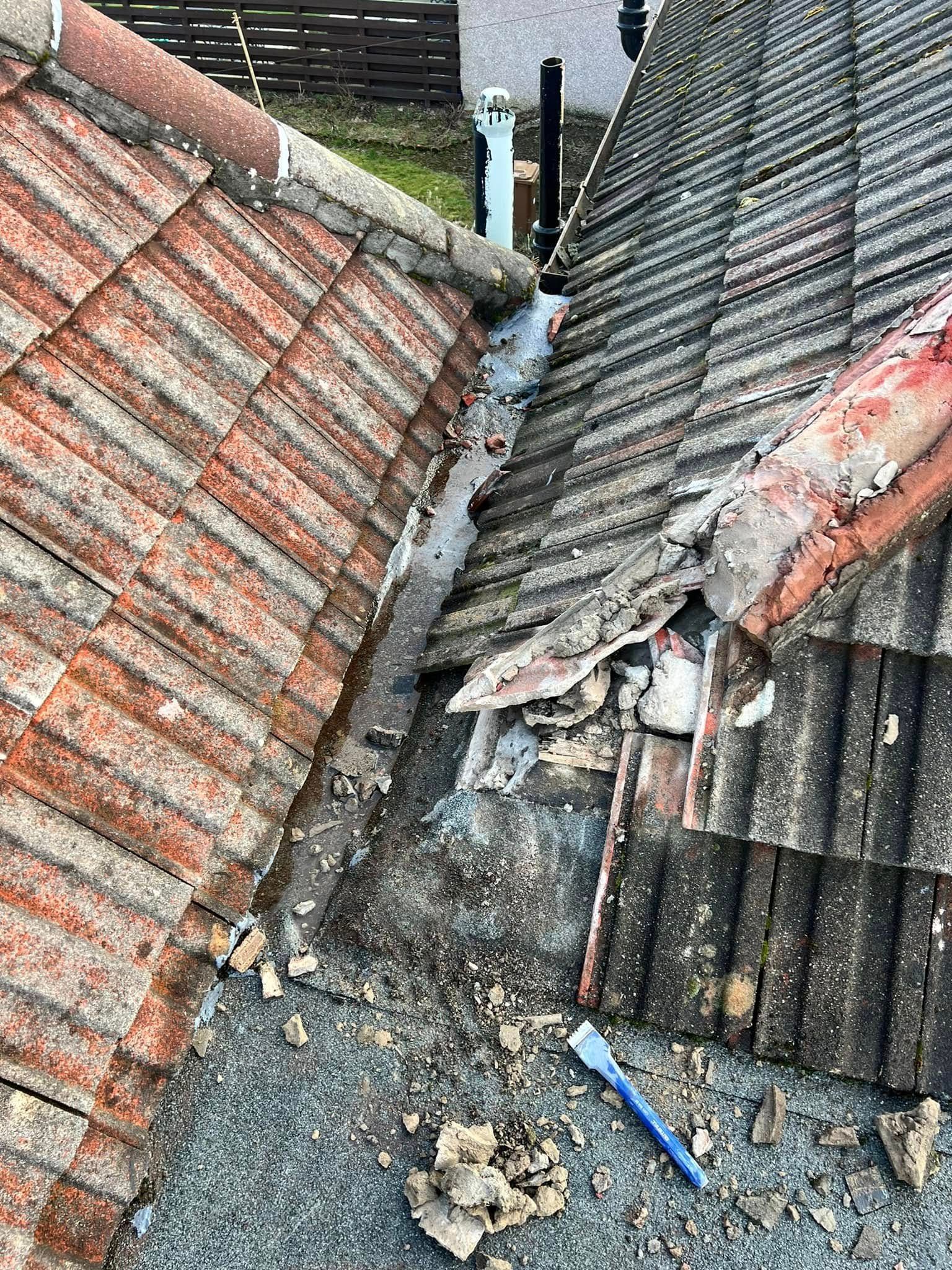 A close-up of a damaged roof valley with cracked and broken tiles, exposed timber and rubble, and a paintbrush lying on the tiles.