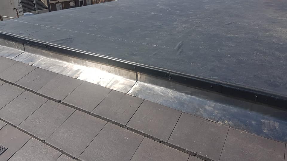 Close-up of a flat rubber roof edge with metal trim meeting a tiled walkway.