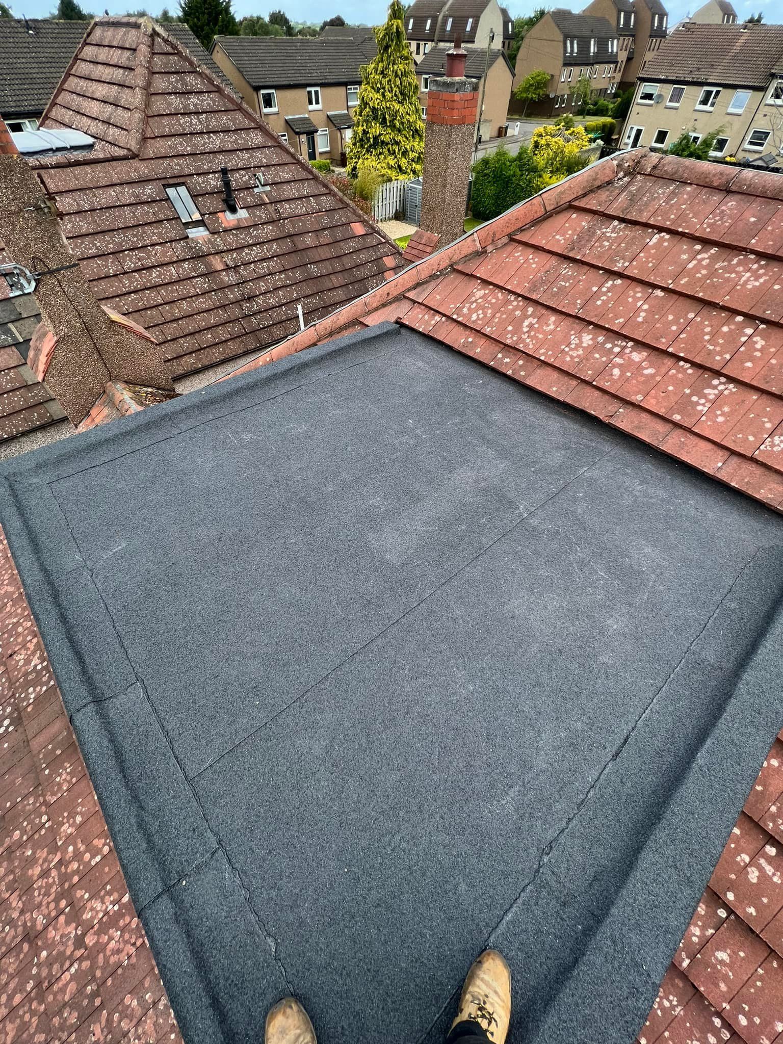 A flat grey felt roof section built between two pitched tile roofs, illustrating how flat and pitched shapes meet on real properties. A flat grey felt roof section built between two pitched tile roofs, illustrating how flat and pitched shapes meet on real properties.