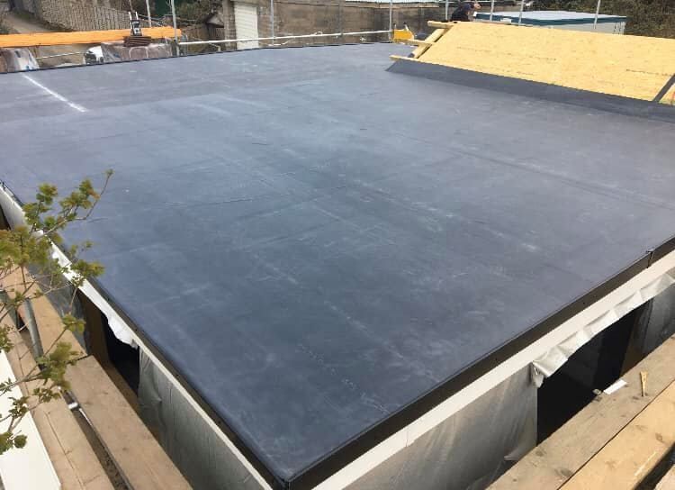 A newly installed grey EPDM rubber membrane on a flat roof extension, with plywood decking and roof tiles visible behind. A newly installed grey EPDM rubber membrane on a flat roof extension, with plywood decking and roof tiles visible behind.