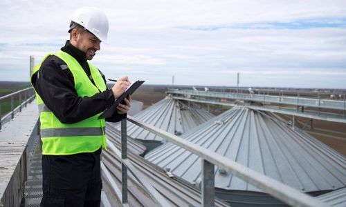 A roofing inspector in a hi-vis vest and hard hat writing on a clipboard on an elevated walkway overlooking corrugated metal silo roofs. A roofing inspector in a hi-vis vest and hard hat writing on a clipboard on an elevated walkway overlooking corrugated metal silo roofs.
