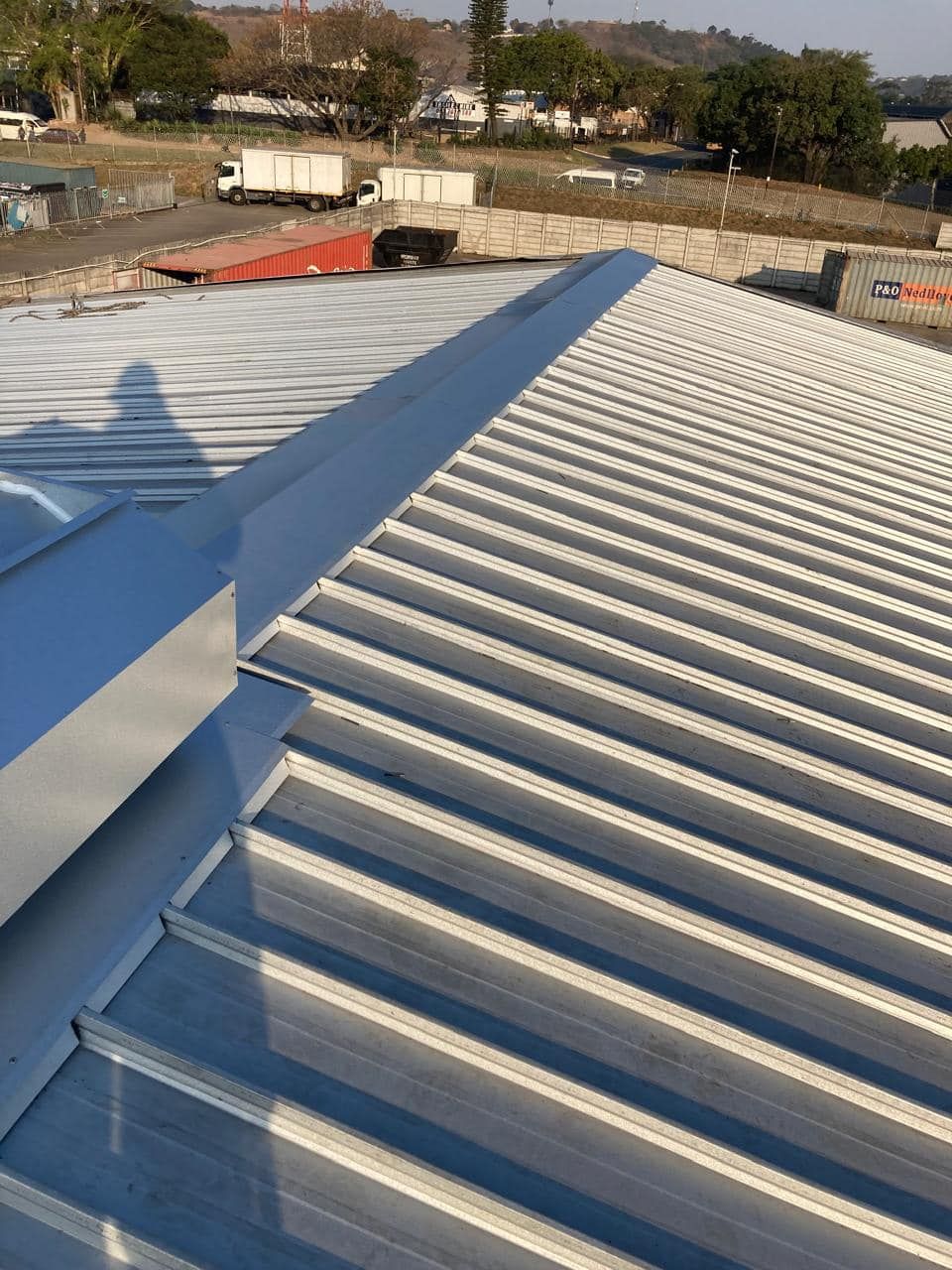 A wide expanse of silver corrugated metal roofing on an industrial building, with a yard, shipping containers, and trees visible in the background. A wide expanse of silver corrugated metal roofing on an industrial building, with a yard, shipping containers, and trees visible in the background.
