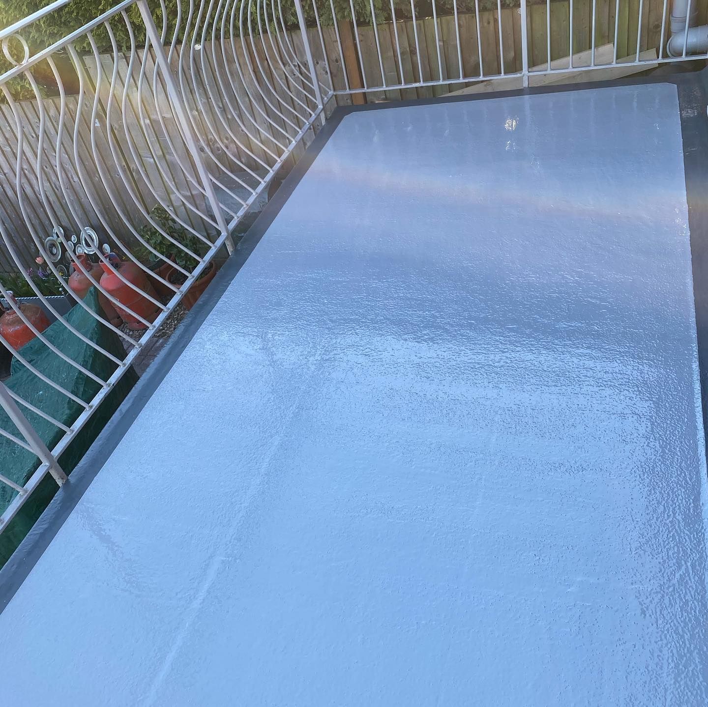 A pale blue-grey GRP fibreglass flat roof forming a balcony area, edged with white ornate metal railings and a wooden fence behind.