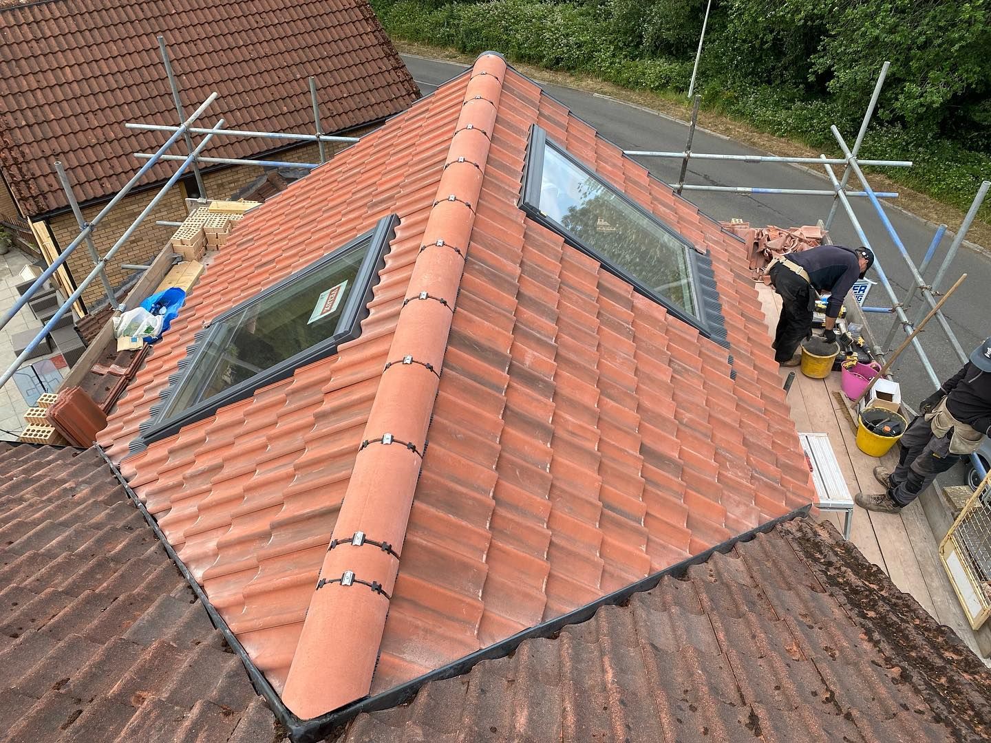 An aerial view of a red tiled roof dormer with two skylight windows, a ridge tile capped with clip fixings, and workers on scaffolding nearby. An aerial view of a red tiled roof dormer with two skylight windows, a ridge tile capped with clip fixings, and workers on scaffolding nearby.