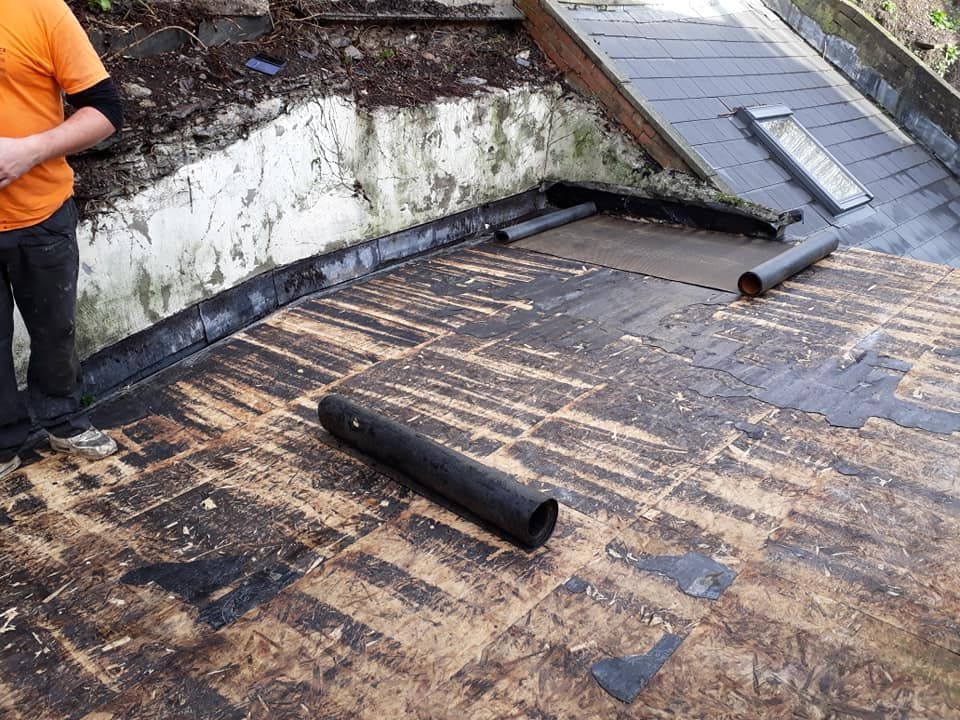 A stripped flat roof deck showing bare plywood boards and rolled bitumen felt, with a worker standing beside the parapet wall.